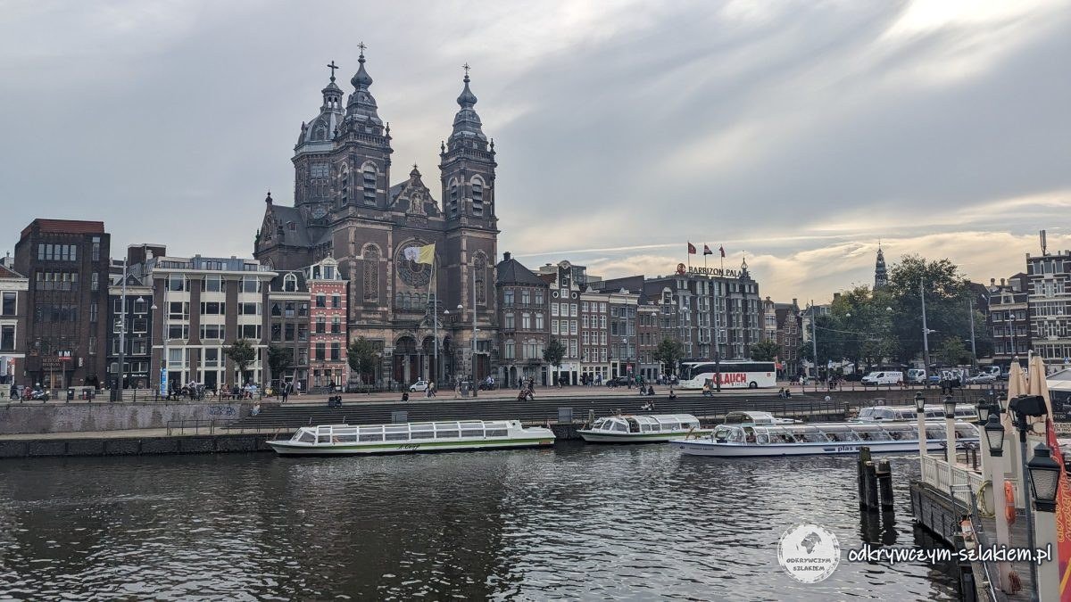 St Nicholas Basilica and waterfront houses in Amsterdam under a cloudy sky, with tour boats in the foreground