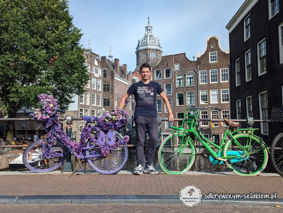 Maciek between a bike covered in purple flowers and a green bike on a canal bridge in Amsterdam, with the basilica dome behind