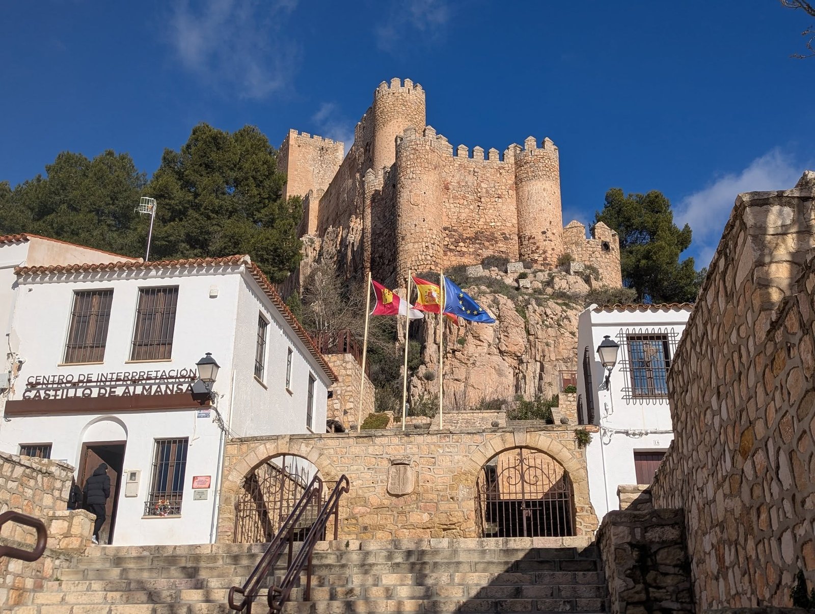 Zamek w Almansie na skale nad schodami i centrum informacyjnym Castillo de Almansa, z flagami Kastylii-La Manchy, Hiszpanii i Unii Europejskiej