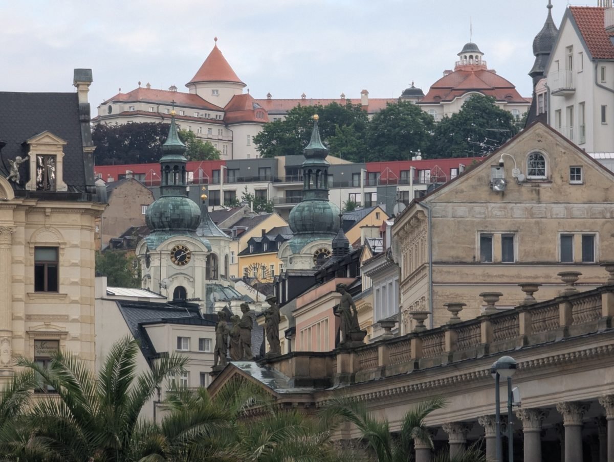Balustrada z rzeźbami na kolumnadzie, a za nią kościół o dwóch wieżach i hotele na wzgórzu w Karlowych Warach