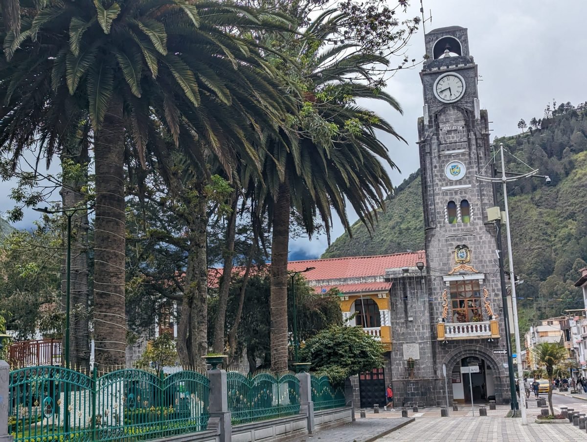Kamienna wieża zegarowa i palmy przy placu w centrum Baños de Agua Santa, w tle zielone zbocze góry