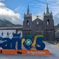 Kolorowy napis Baños de Agua Santa na placu, za nim bazylika z ciemnego kamienia i zielone góry