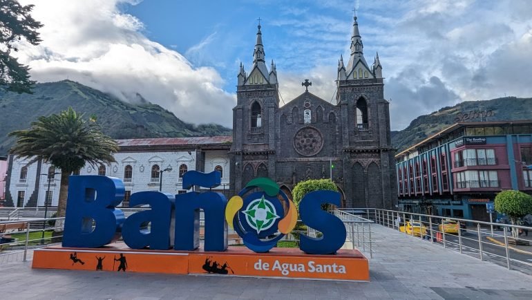 Kolorowy napis Baños de Agua Santa na placu, za nim bazylika z ciemnego kamienia i zielone góry