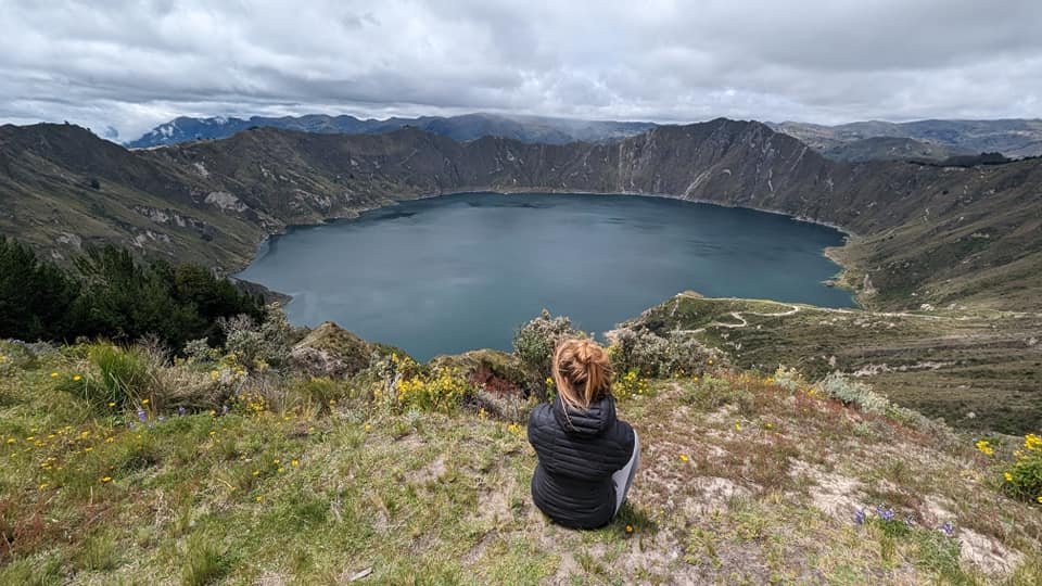Patrycja siedzi na trawie nad laguną Quilotoa, w tle ściany krateru