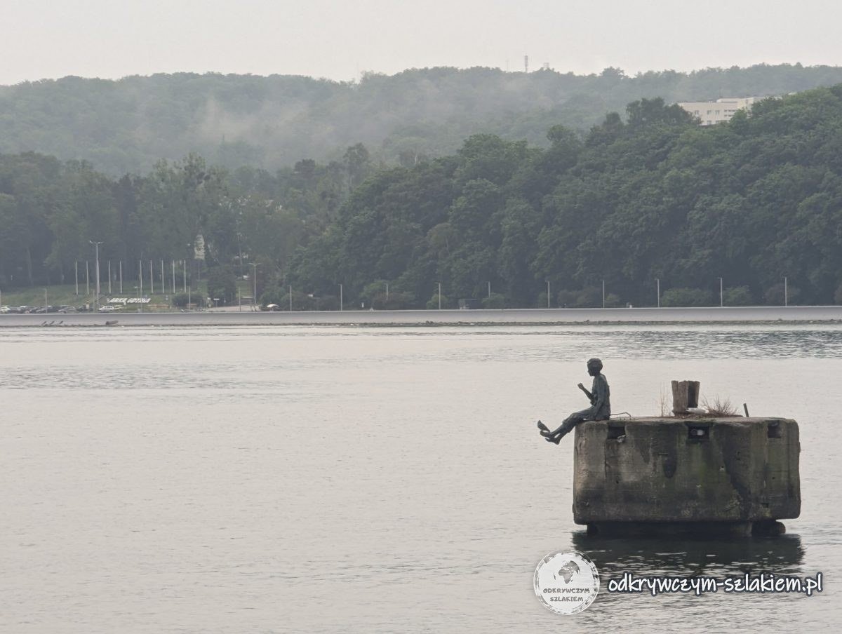 The Dreamer sculpture of a boy on a concrete mooring dolphin in the water by the South Pier in Gdynia, with a wooded hill in the mist behind