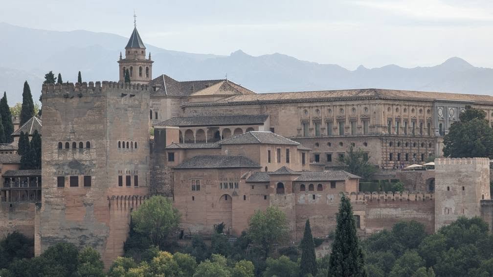 Alhambra w Grenadzie – mury i wieże twierdzy na wzgórzu nad zielenią, widziane od strony dzielnicy Albaicín