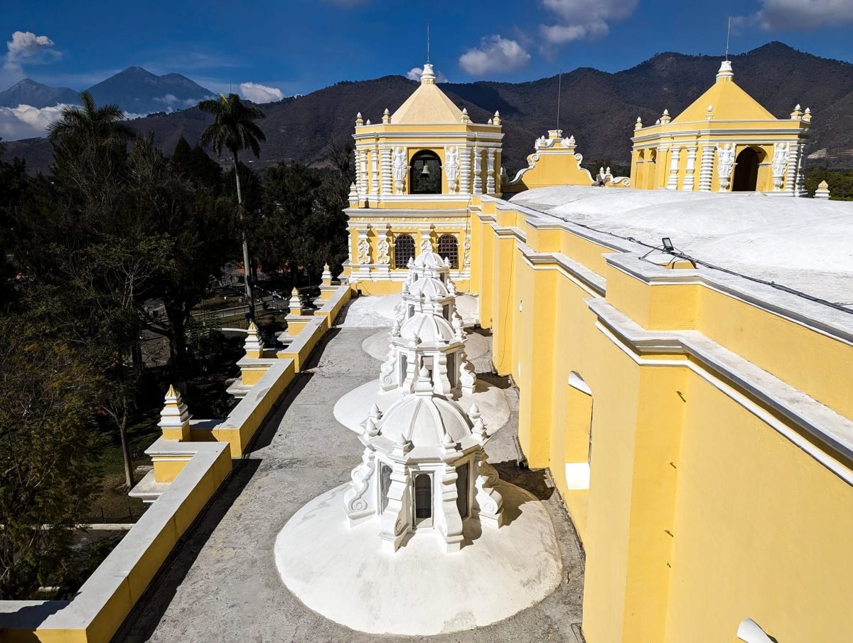 Żółty kościół z dwiema wieżami i biała barokowa konstrukcja przed nim, za nimi góry – Antigua Guatemala