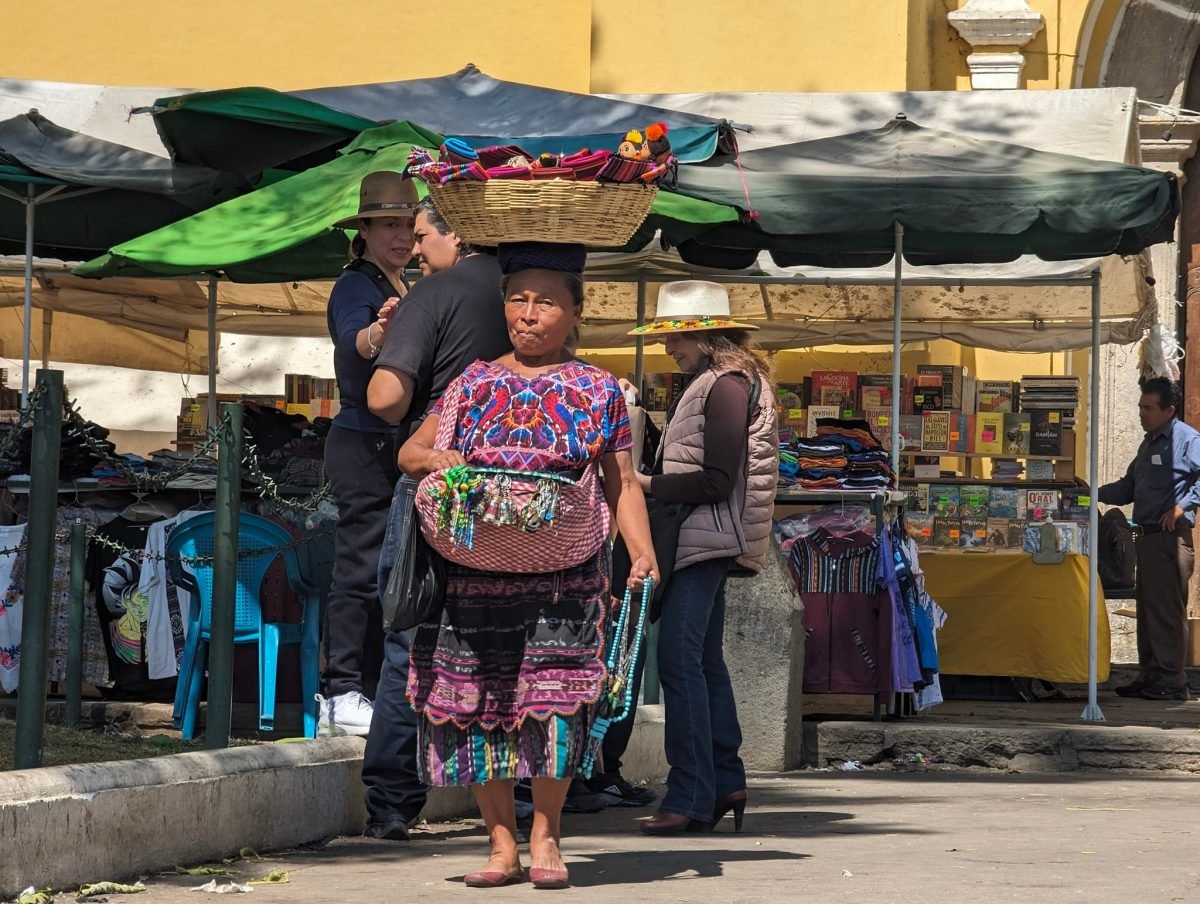 Kobieta w kolorowym tradycyjnym stroju z koszem na głowie przy straganach w Antigua Guatemala