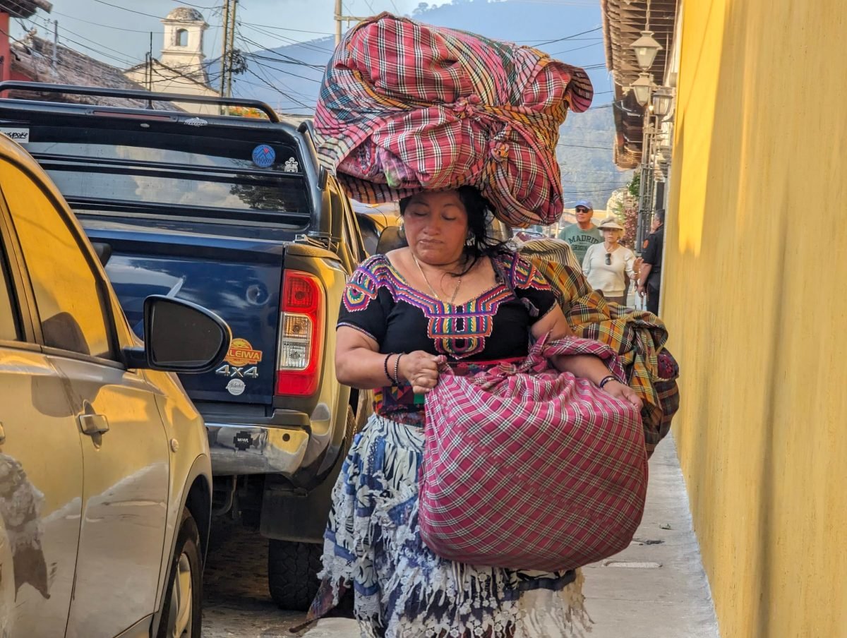 Kobieta w tradycyjnej haftowanej bluzce niesie na głowie wielki tobołek z tkaninami, ulica w Antigua Guatemala