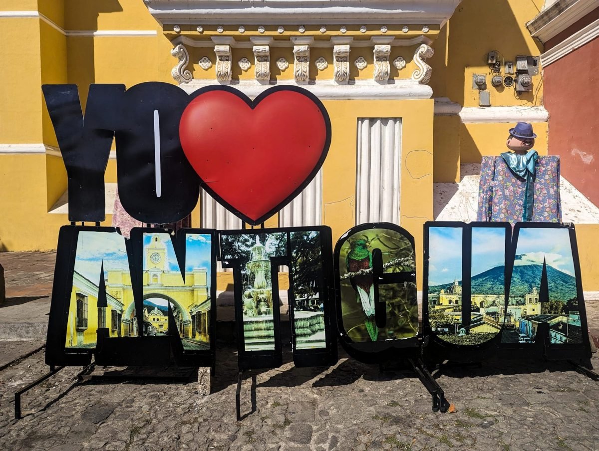 Duży kolorowy napis z czerwonym sercem i słowem „Antigua” przed żółtym budynkiem w Antigua Guatemala