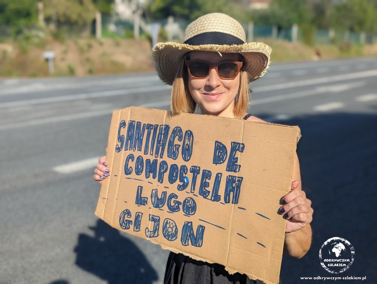Patrycja z tekturową tabliczką „Santiago de Compostela – Lugo – Gijón” przy drodze