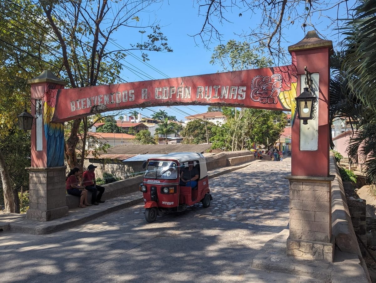 Brama z napisem Bienvenidos a Copán Ruinas i czerwona mototaksówka przy wjeździe do miasteczka