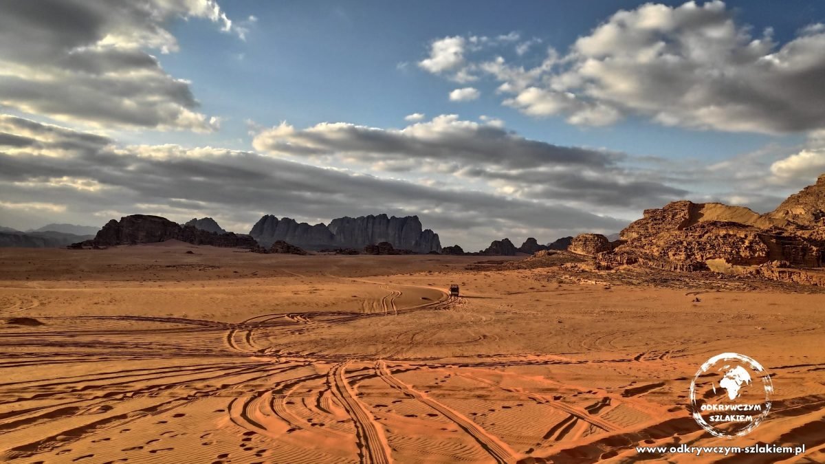 Ślady kół na czerwonym piasku pustyni Wadi Rum, w tle skalne masywy