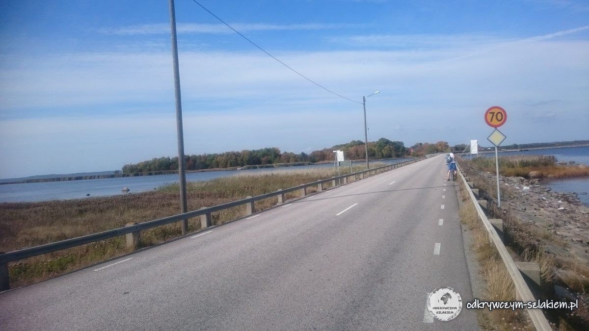 Una carretera asfaltada por un terraplén sobre el agua camino de la isla de Hasslö, con un ciclista a lo lejos y una señal de 70 km/h