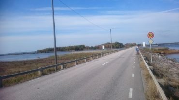 Asfaltowa droga po grobli nad wodą w drodze na wyspę Hasslö, z rowerzystą w oddali i znakiem ograniczenia do 70 km/h