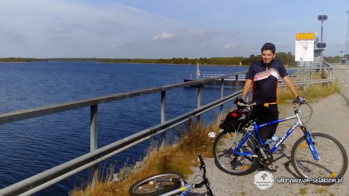 Un ciclista con una bici azul junto a la barandilla del terraplén sobre el agua, cerca del puente de Hasslö