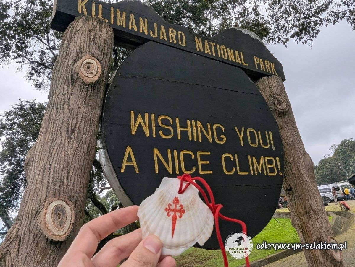 Muszla świętego Jakuba na tle tablicy Wishing you a nice climb przy Machame Gate