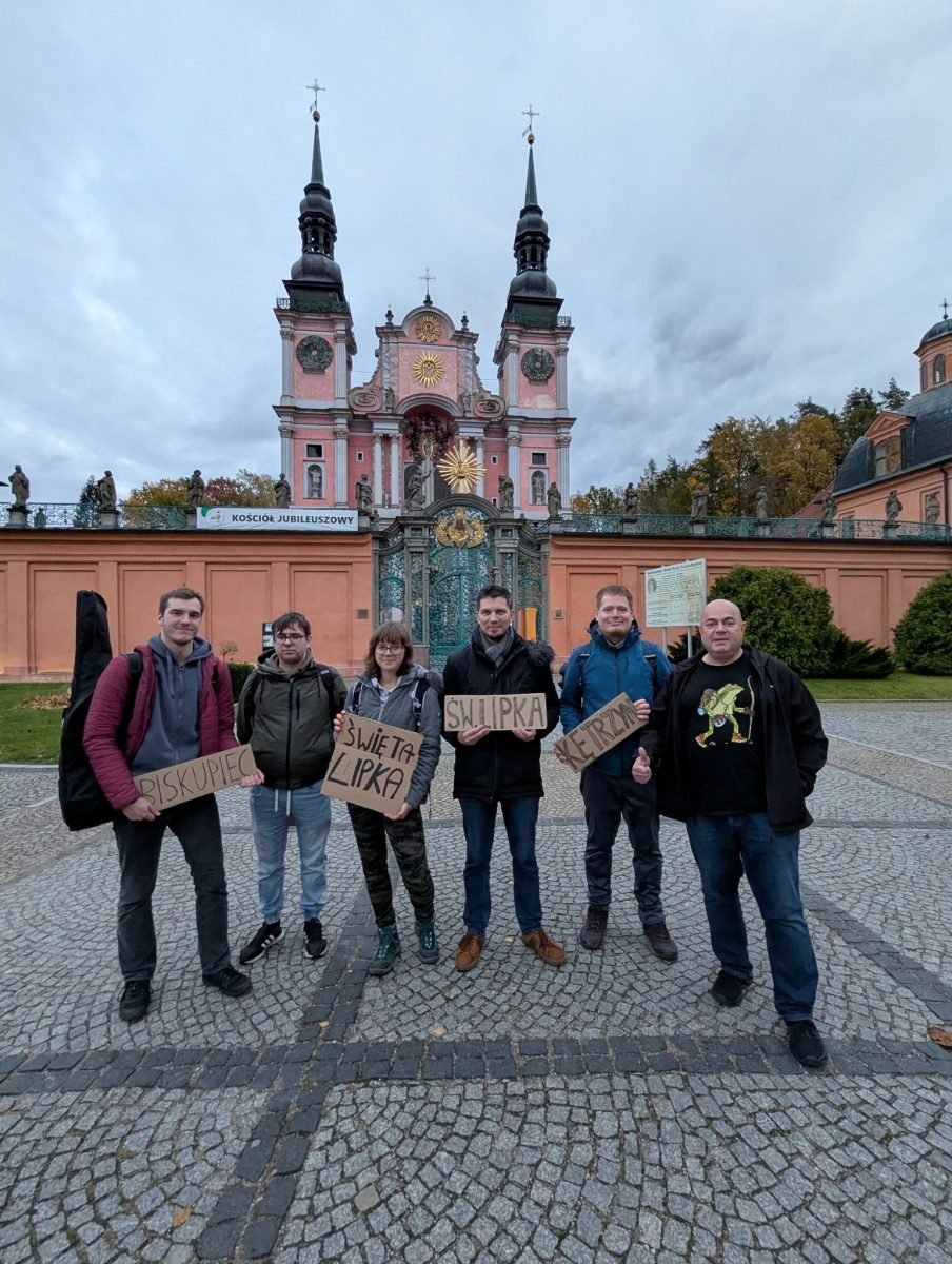 Uczestnicy rajdu autostopowego z kartonowymi tabliczkami przed bramą bazyliki w Świętej Lipce