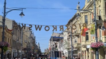 Die Piotrkowska-Straße in Łódź im Sommer – Schriftzug über der Fußgängerzone, bunte Mietshäuser mit Blumen und viele Spaziergänger