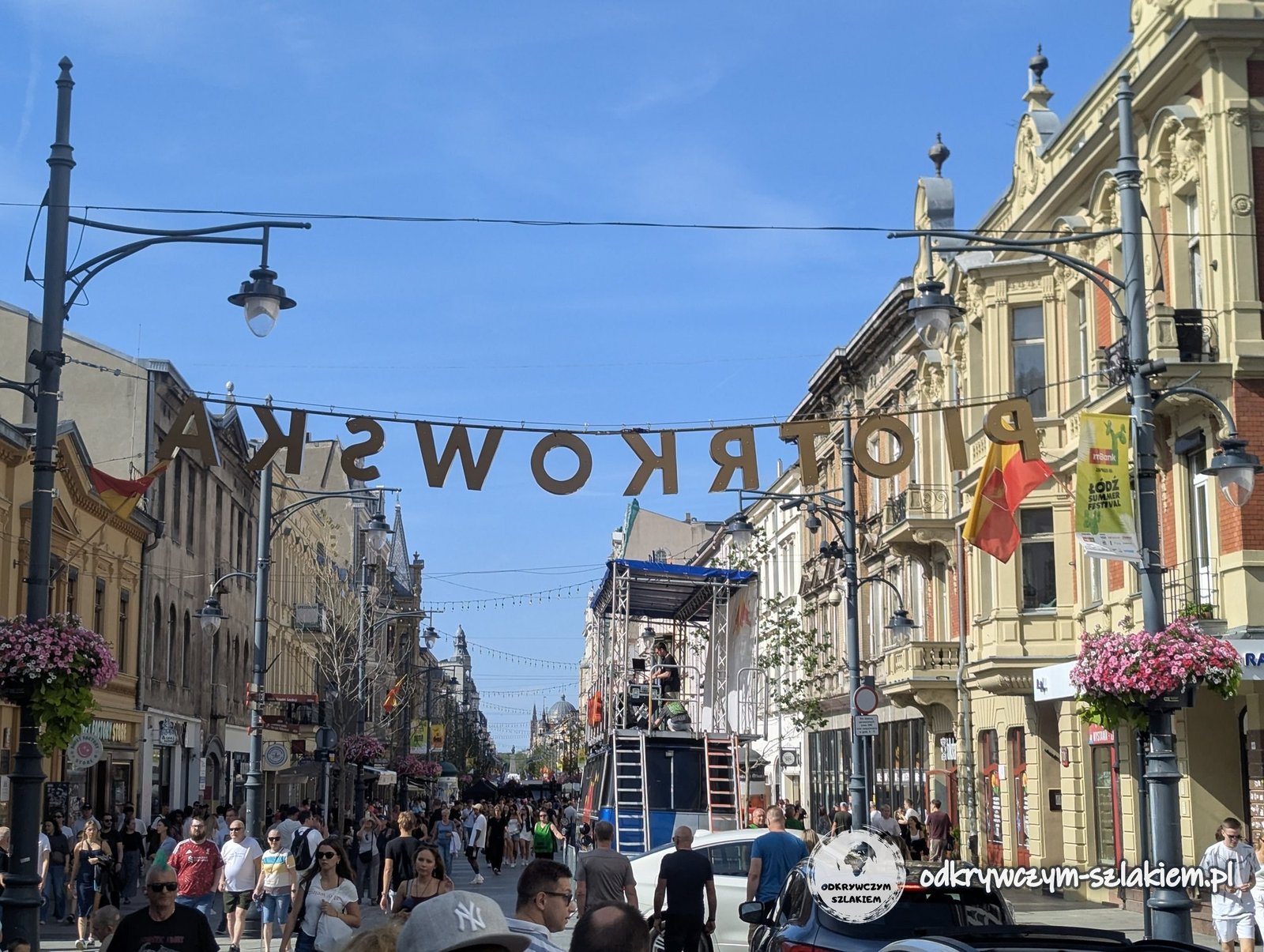 Ulica Piotrkowska w Łodzi latem – napis nad deptakiem, kolorowe kamienice z kwiatami i tłum spacerowiczów