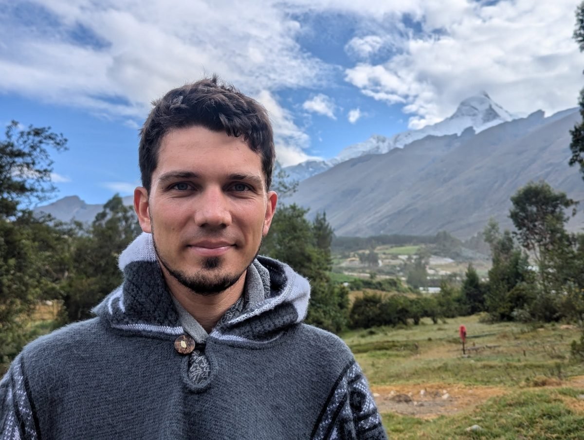 Maciek w swetrze o poranku, za nim łąka, drzewa i ośnieżony szczyt Cordillera Blanca