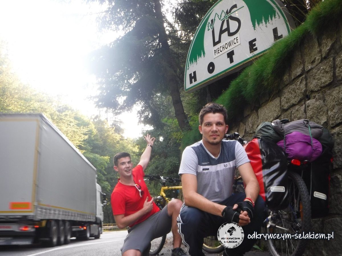 Maciek y Michał con las bicis cargadas junto a un muro de piedra bajo el letrero del hotel Las en Piechowice