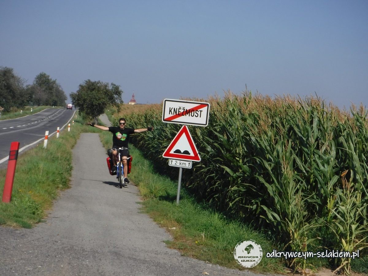 Maciek con los brazos abiertos pedaleando con la bici cargada por un camino junto a un maizal, al lado del cartel de fin de Kněžmost