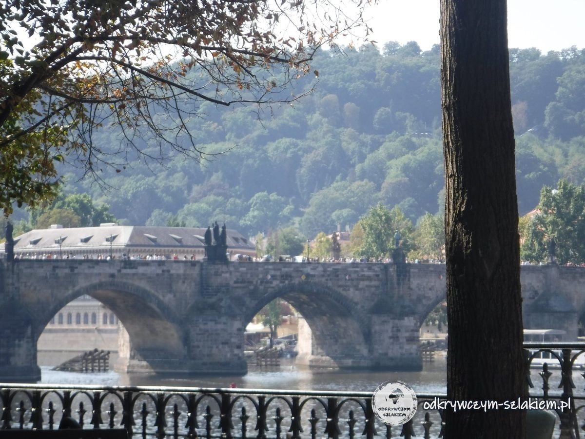 Los arcos del Puente de Carlos vistos tras la barandilla de hierro de la orilla de la Ciudad Vieja, con una colina boscosa detrás