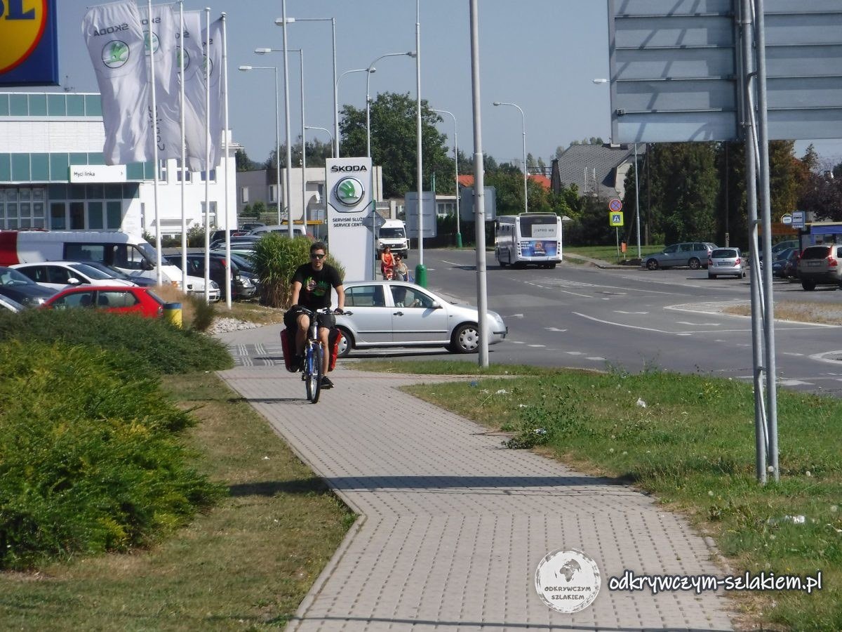 Maciek pedalea con la bici cargada por la acera junto a un concesionario Škoda con banderas en Mladá Boleslav
