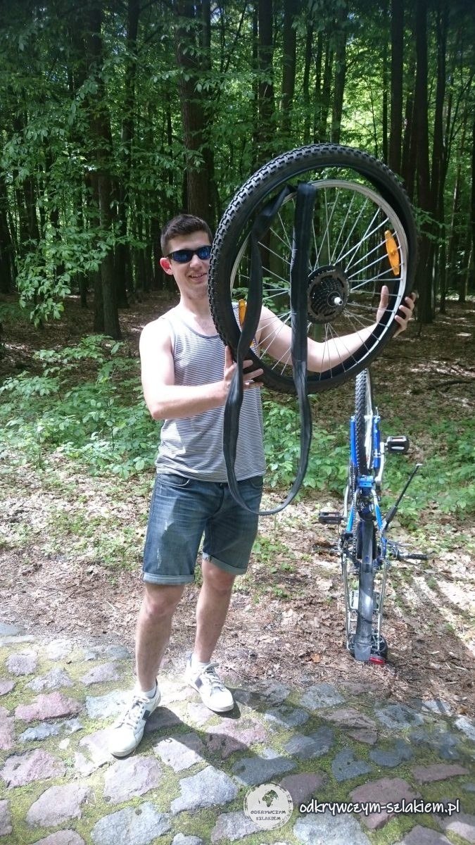 Michał trzyma koło roweru z przebitą dętką na brukowanej drodze w lesie