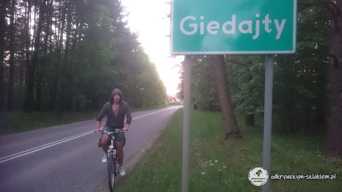 Michał na rowerze przy tablicy z nazwą wsi Giedajty o zmierzchu