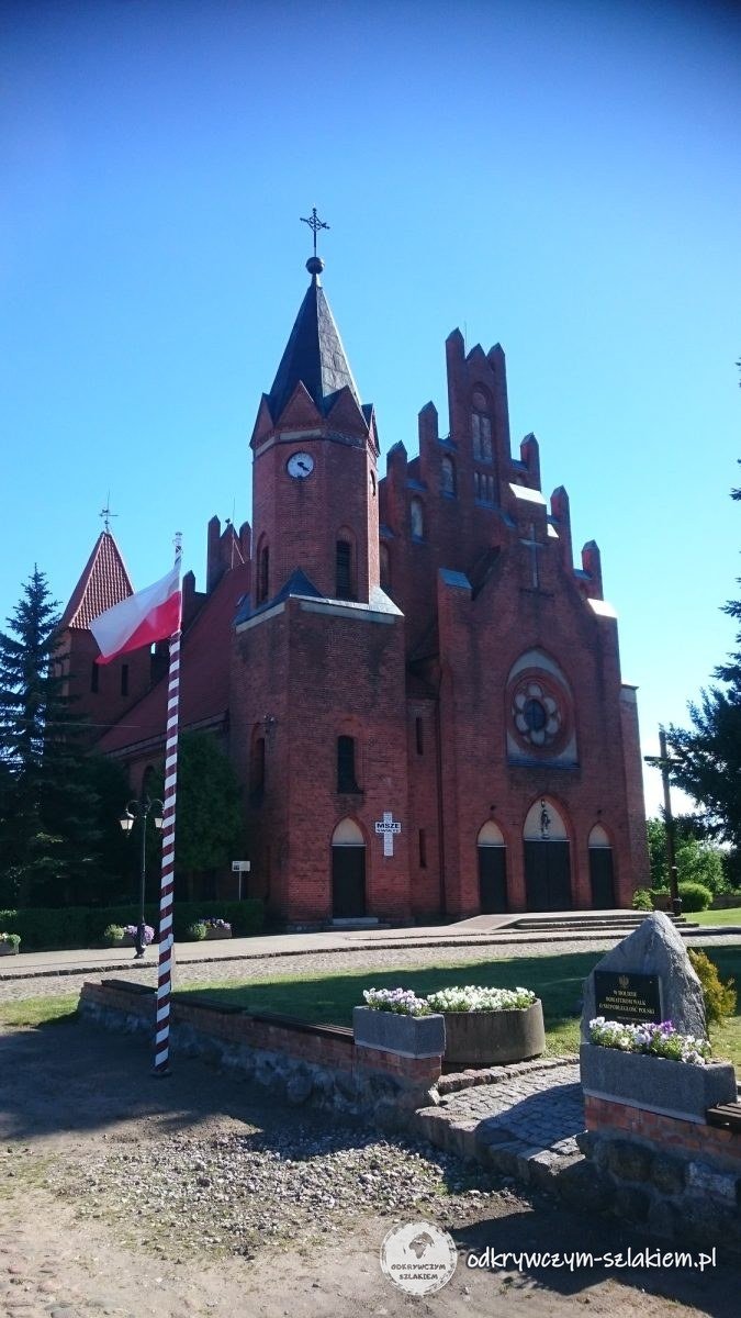 Ceglany kościół z wysoką wieżą i flagą przy drodze na trasie przez Miłomłyn