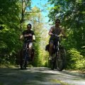 Two cyclists with panniers riding side by side along a sunny forest road on the way from Malbork to Olsztyn