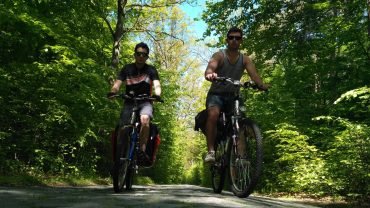 Dwóch rowerzystów z sakwami jedzie obok siebie leśną drogą w słońcu, w drodze z Malborka do Olsztyna