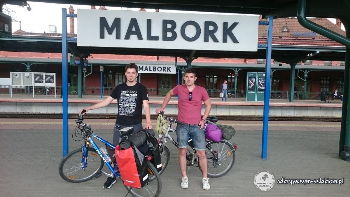Dwóch rowerzystów z rowerami z sakwami pod tablicą z nazwą stacji Malbork