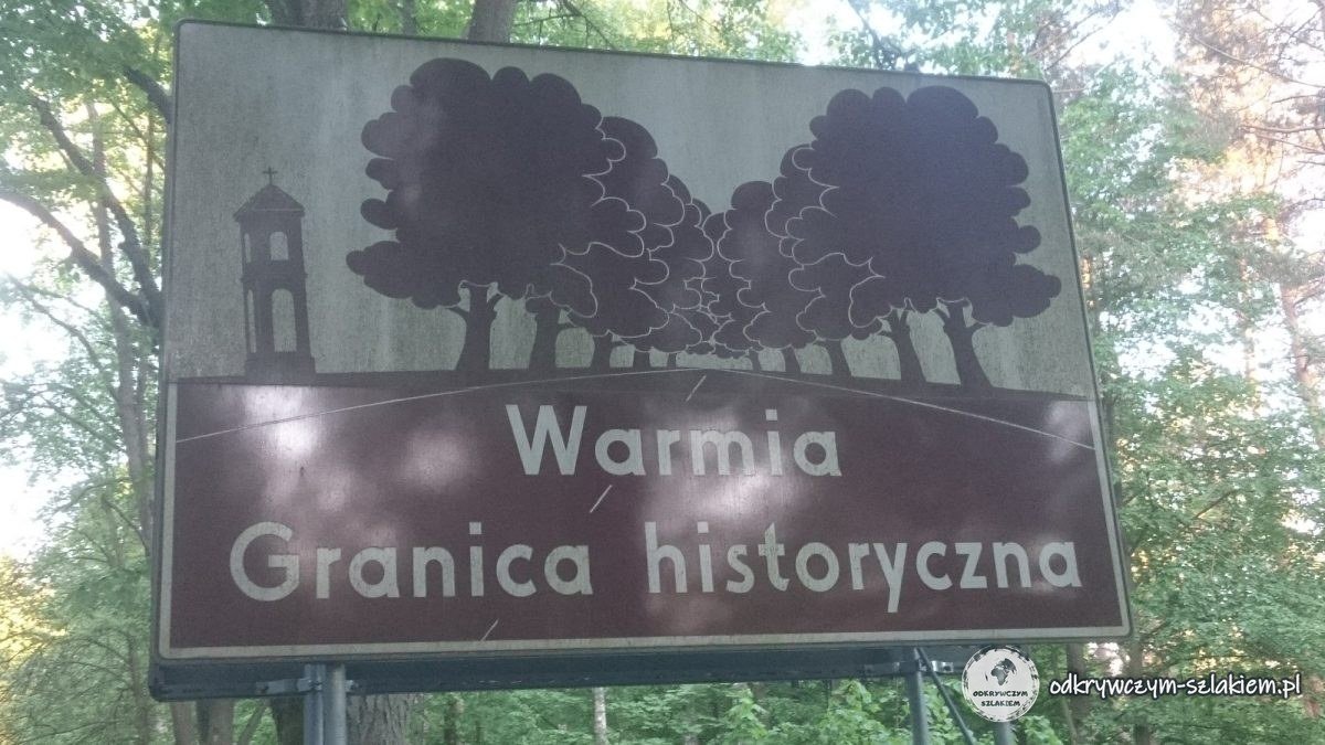 Brązowa tablica „Warmia – granica historyczna” z sylwetką kapliczki i drzew przy drodze z Łukty do Olsztyna