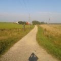 Camino de grava entre prados y campos cerca de Szuszalewo, en el límite del Parque Nacional del Biebrza, con la sombra de un ciclista