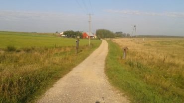 Szutrowa droga między łąkami i polami koło Szuszalewa na skraju Biebrzańskiego Parku Narodowego, z cieniem rowerzysty