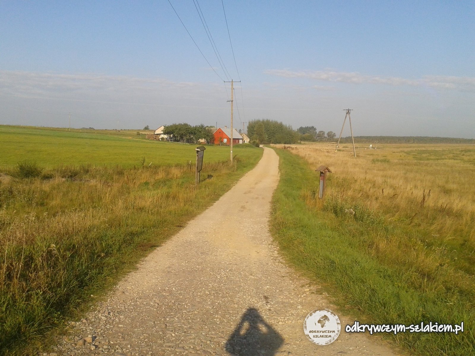 Schotterweg zwischen Wiesen und Feldern bei Szuszalewo am Rand des Biebrza-Nationalparks, mit dem Schatten eines Radfahrers