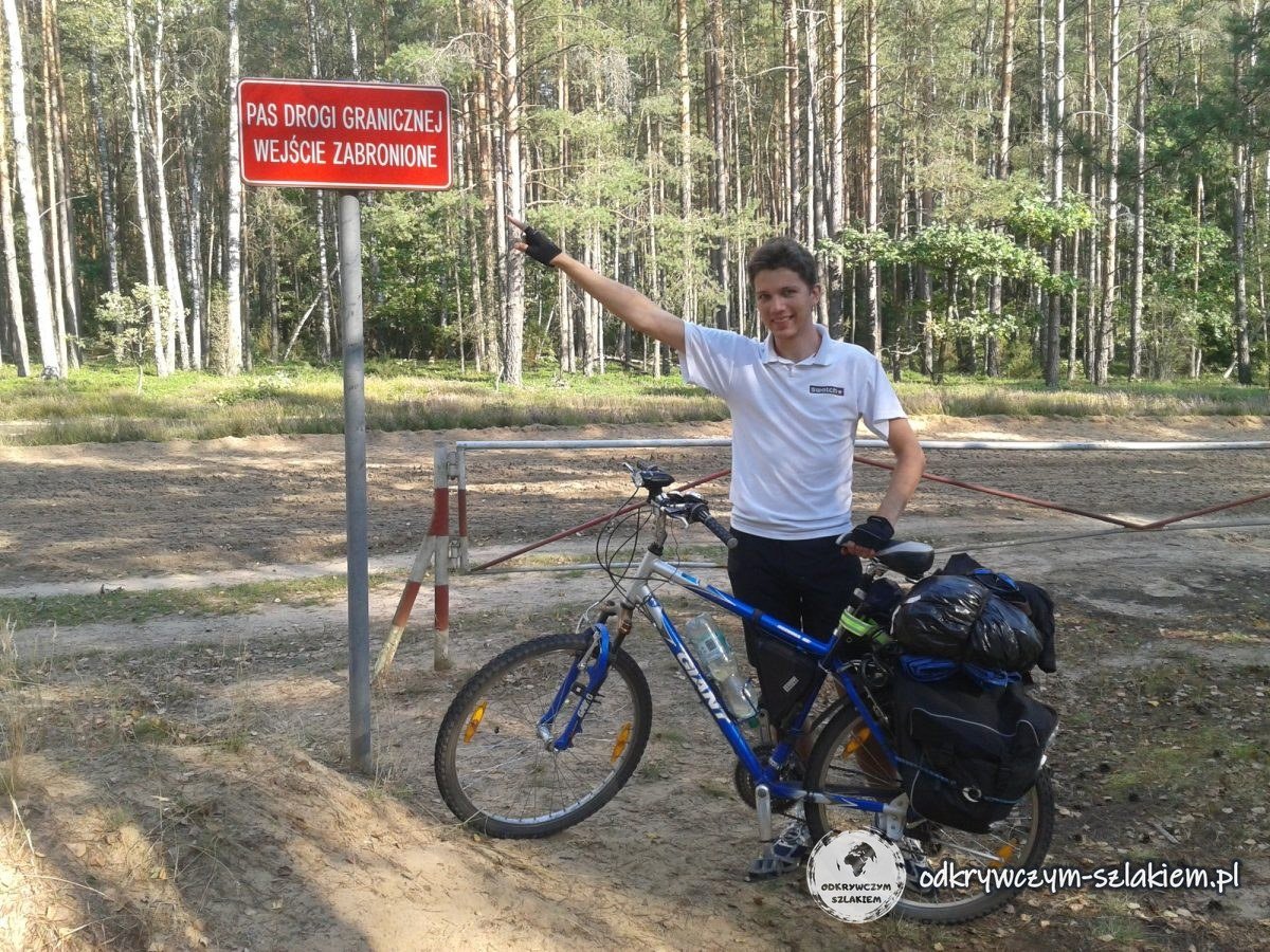 Maciek mit beladenem Rad unter dem Grenzstreifen-Schild an einer Schranke im Wald