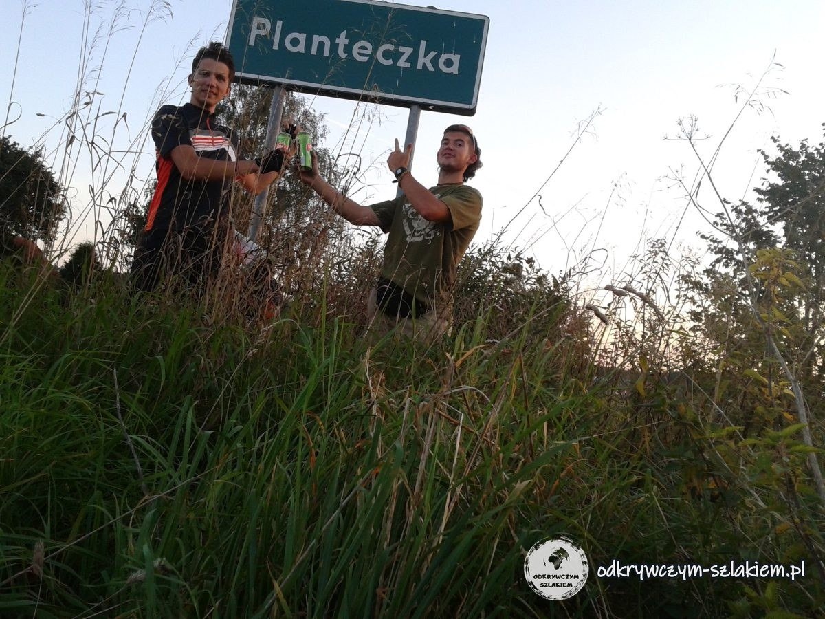 Maciek und Michał am Ortsschild von Planteczka auf einer grasbewachsenen Böschung