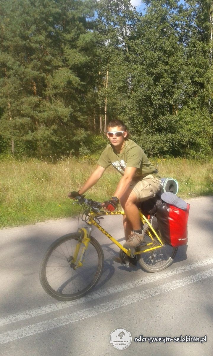 Michał fährt auf einem gelben Rad mit roten Packtaschen eine Asphaltstraße durch den Wald