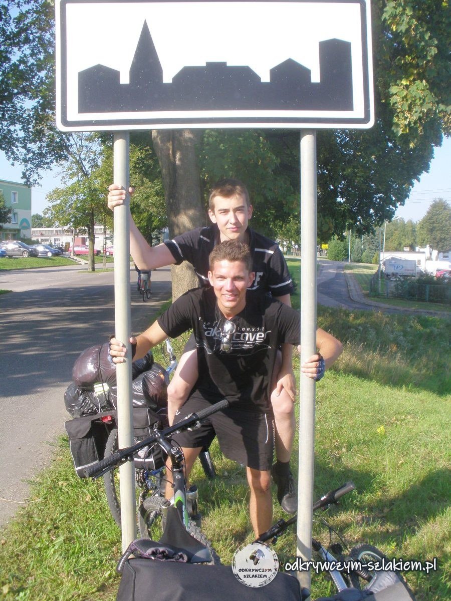 Zwei Radfahrer am Ortsschild von Sensburg (Mrągowo), daneben Räder mit Packtaschen