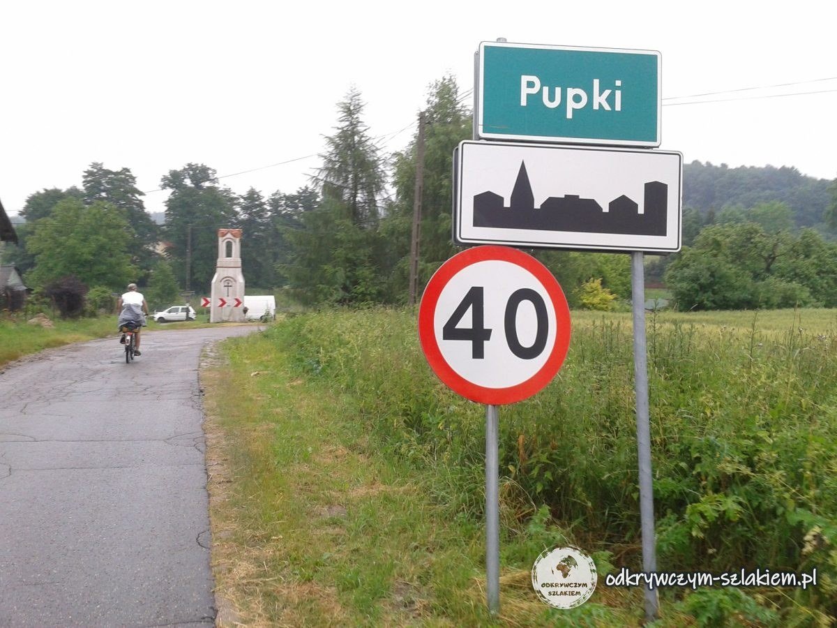 Ortsschild von Pupki, eine Kapelle und ein Radfahrer auf schmaler Straße an einem trüben Tag