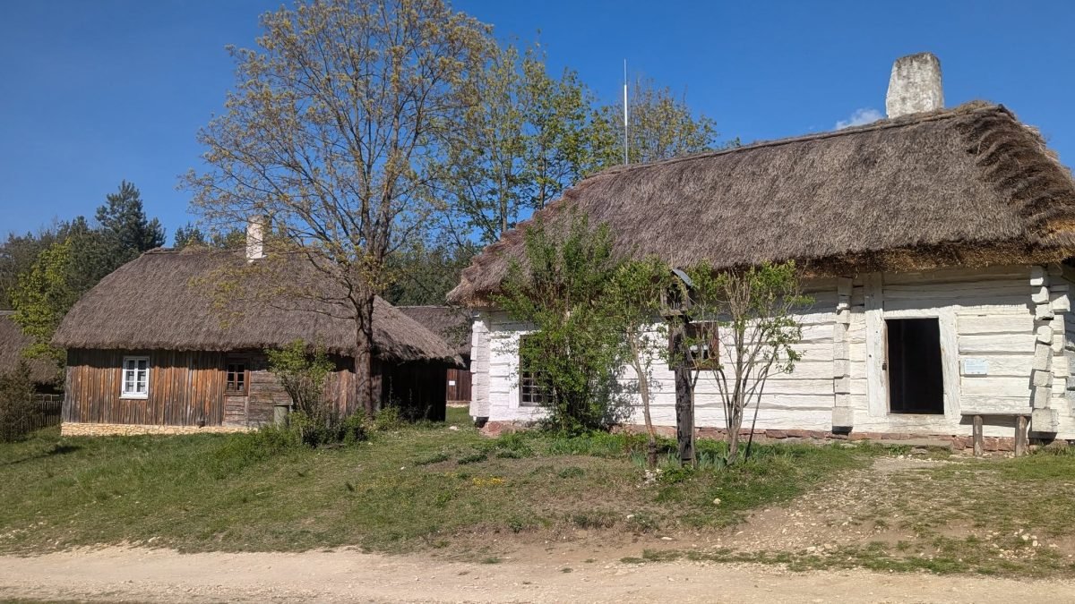 Dwie chaty kryte strzechą wśród drzew w skansenie w Tokarni
