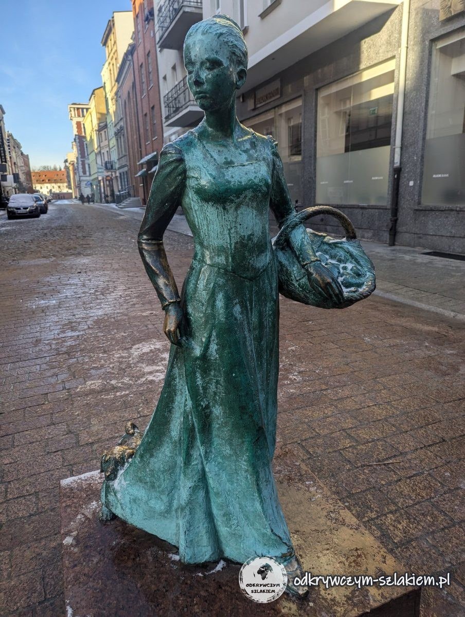 Escultura de la Vendedora de Pan de Jengibre de Toruń con una cesta en una calle del casco antiguo