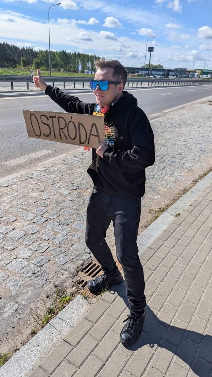 Krzysiek łapie stopa z tabliczką „Ostróda” na poboczu drogi