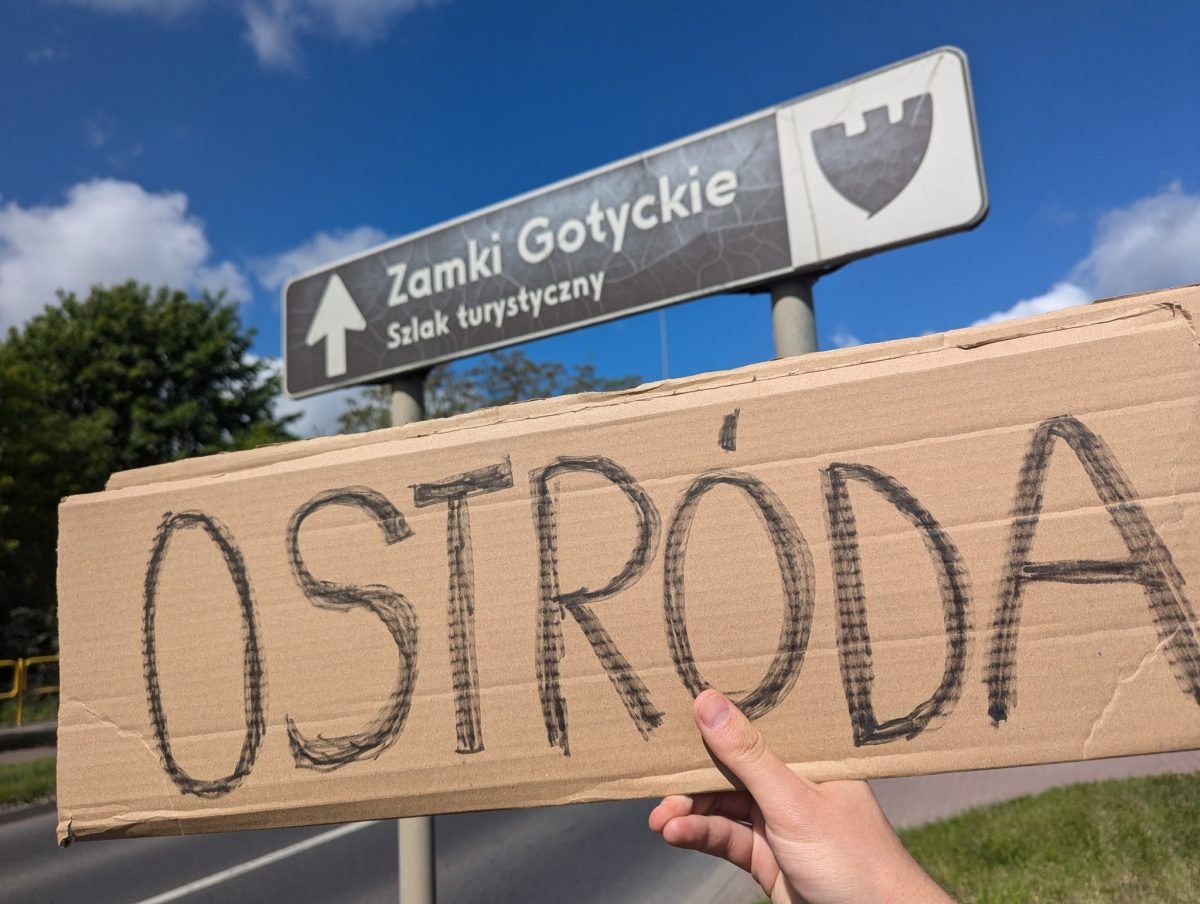 Tabliczka „Ostróda” trzymana pod drogowskazem szlaku turystycznego Zamki Gotyckie