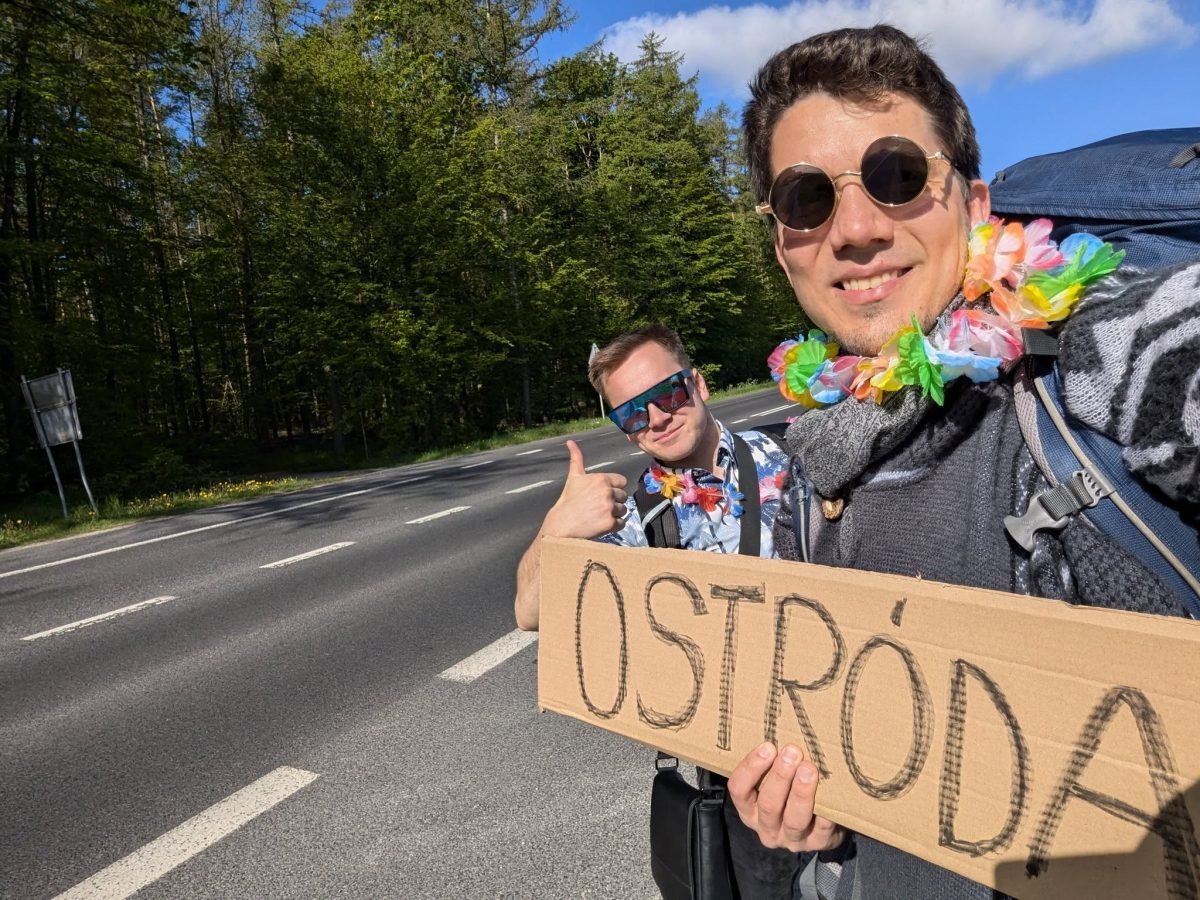 Maciek i Krzysiek z tabliczką „Ostróda” łapią stopa przy drodze przez las