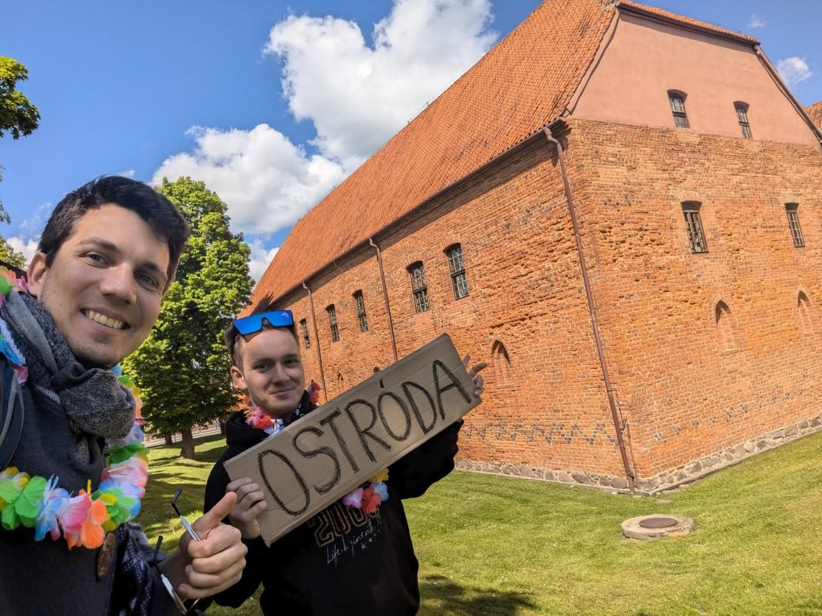 Maciek i Krzysiek z tabliczką „Ostróda” przed ceglanym zamkiem krzyżackim w Ostródzie